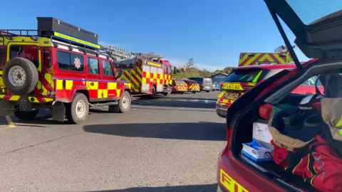 A number of fire trucks and cars lined up together. The sky is blue above. 