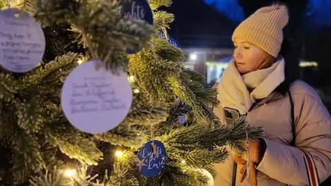 A woman looking towards a Christmas tree which features lights and cardboard baubles with dedications.