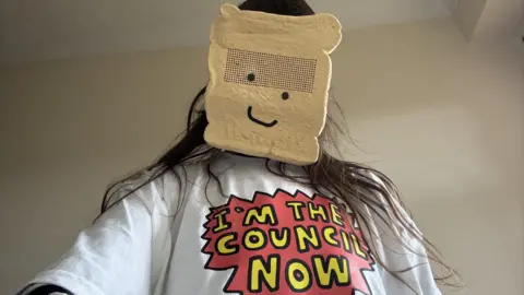 A person with long brown hair poses for a selfie, with a mask covering their face. The mask looks like a slice of white bread, with two black dots for eyes and a black line in the shape of a wonky smile. They are also wearing a white t-shirt with a slogan in yellow writing on a red background, which reads: "I'm the council now"
