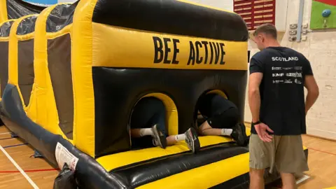A large black and yellow inflatable obstacle course with the words Bee Active on it. The feet of two people climbing into the inflatable can be seen, and another man is looking on