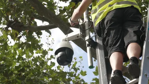 Picture of a CCTV camera being installed by a man wearing a hi-viz jacket on a ladder next to a lamp post