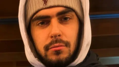 Man looking at the camera he is wearing a woolly hat and a hooded top. He has a short brown beard and moustache.