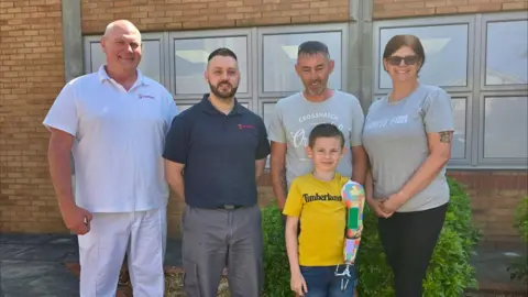 Portsmouth Hospitals University NHS Trust Three men, a woman and a young boy with a prosthetic arm standing in front of a building and smiling at the camera.