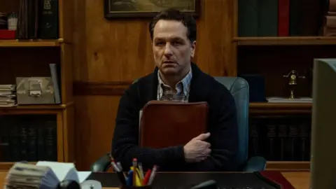 Apple TV Matthew Rhys in Widow's Bay. He is sat down, holding a folder, looking straight ahead 