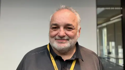 Shariqua Ahmed/BBC Head and shoulders image of Christian Hogg. He has short grey hair, a grey beard and is wearing a dark grey polo shirt with a yellow lanyard.