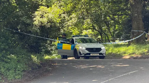 A police car and a police officer cordoned off a part of a park.