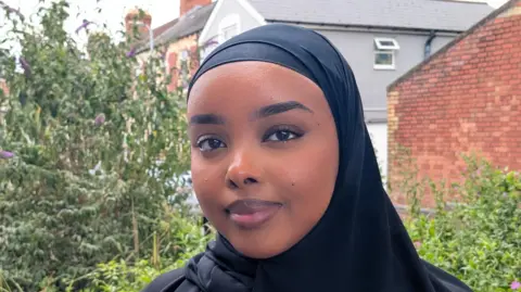 Munirah, a young Welsh-Somali woman stands on a street in Grangetown, Cardiff with trees and houses behind her. She's looking at the camera, smiling and wearing a black headscarf. 