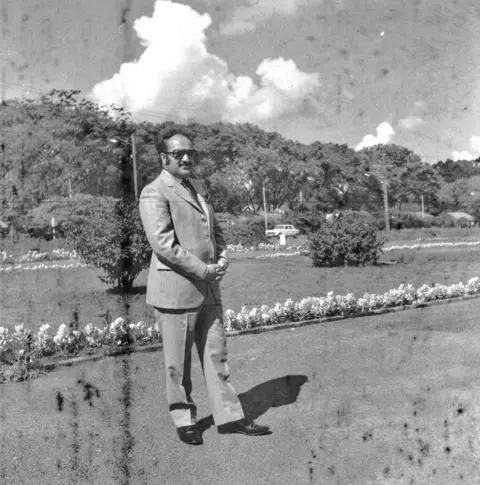 Supplied A black-and-white photograph of Susheel in a smart suit. He has a moustache and is wearing sunglasses. There is a garden with flowers and trees behind him.