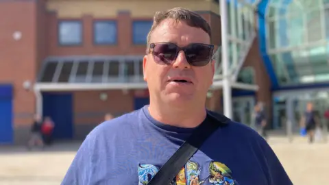 A man with short brown hair and sunglasses. He wears a navy T-shirt and is stood in front of a shopping centre.