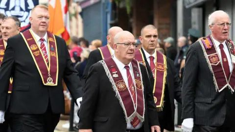 Pacemaker A number of men wearing suits with red ties, and ceremonial garments, marching in a parade.