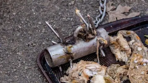 A pile of wet wipes that have been dredged up from a sewage pipe. They are filthy and discoloured. Next to them is the so-called 'baby hedgehog' tool, a small metal cylinder with several spiky nails protruding from various points. It is connected to a metal chain. 