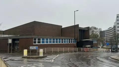 A wider view of the former crown court in Ipswich. A roundabout can be seen in front of the building. Other large office block buildings can be seen in the distance on the right. 