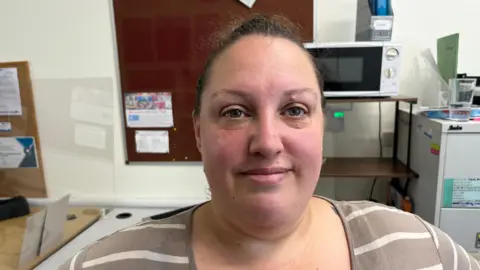 John Devine/BBC Jodie Montgomery, 40, has black high pulled tightly over her head, she ahs a light brown and crème striped jersey on and behind her are items in an office, a noticeboard and a microwave and part of a tall filling cabinet.