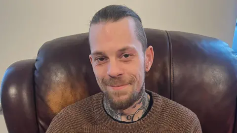 Curtis Durose sitting on a brown leather armchair. Has has brown hair tied back, brown eyes, wearing a brown jumper. He has tattoos on his neck.