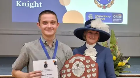 A young man with short dark hair holding a white sheet of paper with writing on it, beside a woman with a blue coat and had holding a wooden shield with metal plates on it
