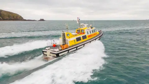 Gaz Papworth Marine ambulance speeding through the sea. The boat is yellow, white and black. Guernsey and ambulance are written in red and its name Flying Christine III is written in white.