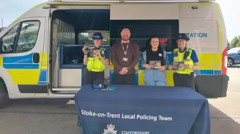 Stoke-on-Trent City Council Two police officers stand next to two people in plain clothing. The four of them are stood behind a table with a blue Staffordshire Police table cloth on it. Behind them is a large white police van with its side door partially open.