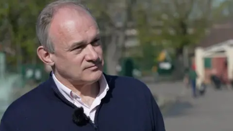 Sir Ed Davey. He is wearing a pink shirt and a dark blue jumper.