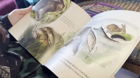 A children's story book being held open above a rattan coffee table. The water colour style illustrations on both pages are of hares. Only a few words of the story are visible, the rest are out of focus. 