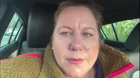 A photo of Kate Jenkins sat in her car. She is looking straight at the phone/camera and is holding it up to her face. She is wearing a brown jumper which has a pink stripe on it.