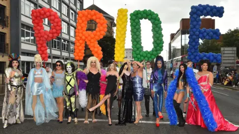 PA Media Drag queens stand in a line in a street in Belfast city centre holding a large balloon sign saying PRIDE. The balloons are in rainbow colours.