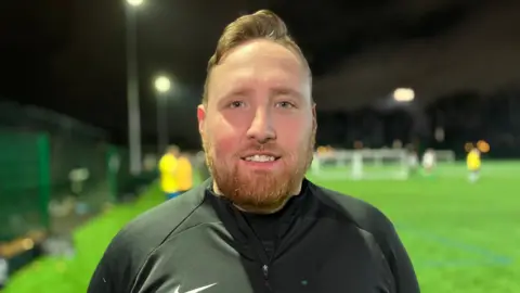 Football player, Lee Simpson, looks into the camera while stood on the sideline at a match