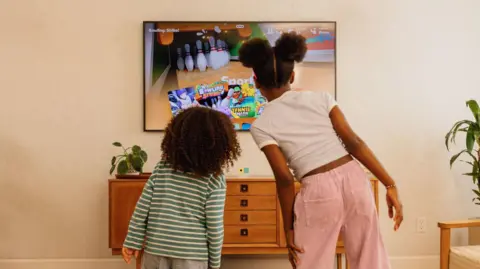 Nex Playground Two children are staring with their backs to the camera in a living room space facing the TV screen which is showing a bowling game. Beneath the screen the Nex Playground console cube in white, green and yellow sits on a table.