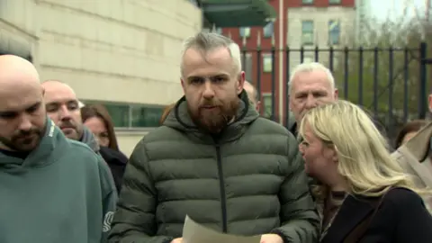 Niall McNally reading a statement outside court. He is wearing a green puffer jacket. He has grey hair and a brown beard.