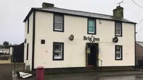 The Brig Inn Pub & Hub The front of the Brig Inn pub, painted white with black paint framing the front door and five windows. A red bin sits to the front left hand side of the building