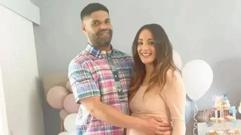 Calvin Buckley holds his arm out over his partner Frankie Jules-Hough as the pair smile for a photograph in front of a set of balloons indoors.