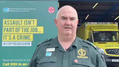 NIAS A man in  green ambulance uniform stands in front of a poster against the abuse of emergency staff. Behind him is an ambulance with florescent livery.