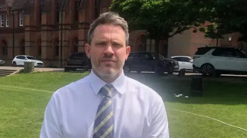 Chris is standing in front of the school building on a grass field.
He is wearing a light coloured shirt and striped tie.
There are trees and cars in the background.
The day is bright and warm.
