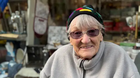 An elderly woman wearing a grey fleece, sunglasses, and a Christmas hat smiling into the camera. A shop front is behind her.