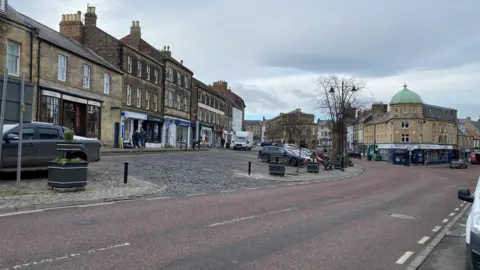 Alnwick town centre. A road sweeps to the left with a turning to the right. Shops sit on either side with an extensive area of cobbles to the left. There are many parked vehicles but only a few people walking around. The sky is cloudy and a little overcast, while a couple of trees have no leaves.