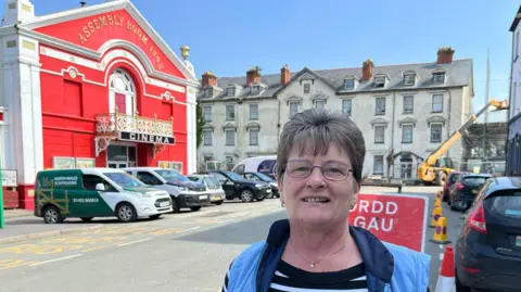 Catherine Evans standing on the road in front of the Corbett Arms Hotel. She has short grey hair and glasses and is wearing a black and white striped shirt with a blue gilet.