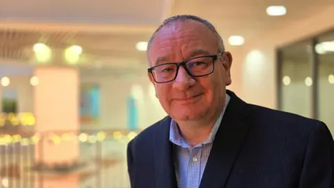 Paul Moseley/BBC Rob Bradley is wearing a checked blue and white shirt beneath a navy suit jacket. He's also wearing glasses. Behind him we can a pillar, railings and various lights in the shopping centre he manages.