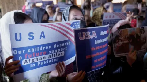 A small crowd of Afghan women carrying signs "The United States of America, our children deserve a future. Support of Afghan allies in Pakistan." Another carries a picture of Donald Trump at the Oval Office. 