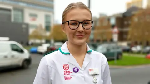 Jenna Watt is wearing a Queens University Belfast white nurse uniform and smiling outside hospital. She has tied up blonde hair and is wearing dark framed glasses. 