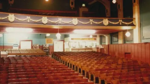 Theatres Trust The inside of a theatre which has rows of seats and a balcony which has a gold design along the front of it. 
