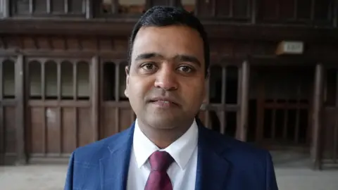 Qays Najm/BBC Dr Sachin Karale smiles at the camera while standing inside County Hall. He has dark, short hair and wears a navy blue suit and a white shirt and red talk. 