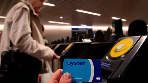 Getty Images Older woman in light brown jacket stands at barriers to London's tube network. A blue Oyster card is held by a hand at the forefront of the shot.