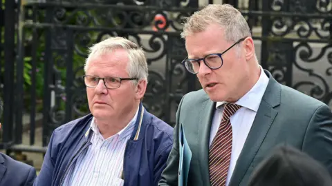 Pacemaker Three people stand behind two different microphones. The man on the right is grey-haired with blue glasses. The man wears a grey suit, white shirt and Brown striped tie. To his right is a grey haired man in black glasses, wearing a blue jacket and a blue and white striped shirt. To the men's right is a man in a navy suit and white shirt, has short brown hair and glasses. T