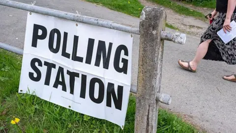 A polling station in Shropshire. There is a metal gate with a paper sign that says POLLING STATION. Someone can be seen walking past with a ballot paper in the background 