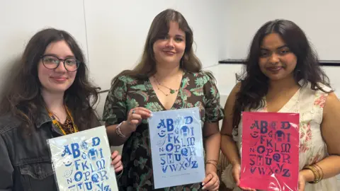 Three young women standing next to each other in front of a white background. They are holding posters with the alphabet printed on them in different fonts.