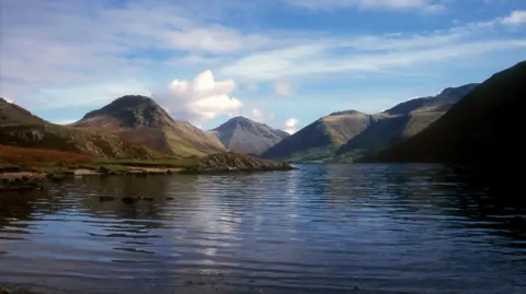 A view of Wast Water in the Lake District taken in 2004. The lake is in the foreground, with small fells on the opposite side of the lake to where the photographer is shooting. The sky is blue with white fluffy clouds. The entire scene is just nature, there are no people, roads or cars.