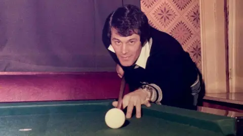 Smith Family Rab Smith playing snooker. He has brown hair and is wearing a dark jumper. He is about to strike the white cue ball with his cue.
