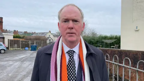 A middle-aged man with short greying hair looks at the camera. He is wearing an orange, white and pink scarf, a white shirt, a dark tie with white spots and a navy blue coat.