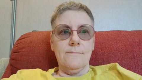 Woman with cropped grey hair round red rimmed glasses yellow t shirt sat on red sofa 