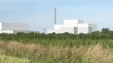 MVV Environment An artist's impression of the new incinerator shows square-shaped white buildings and a large chimney against blue sky. There is a grassed area in the forefront of the image