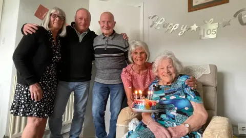 A group of five people are surrounding a birthday cake which has several burning candles on it. You can see Happy 105th birthday on the wall.